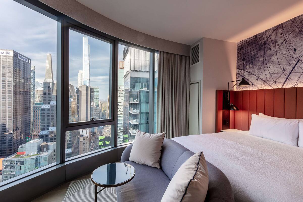 A modern hotel room with a large window offering a city view, a comfortable sofa, a neatly made bed, and a small table.