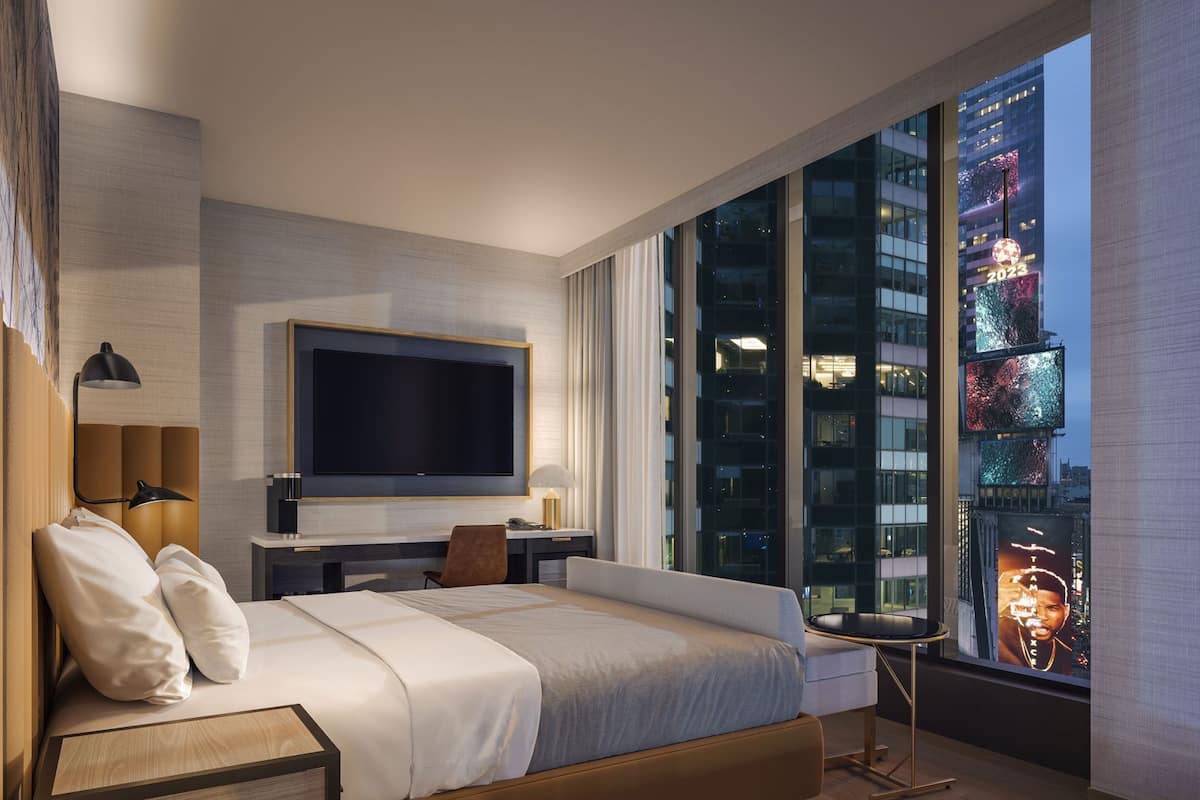 A modern hotel room with a large bed, a flat-screen TV, a desk, and a view of a cityscape at night.