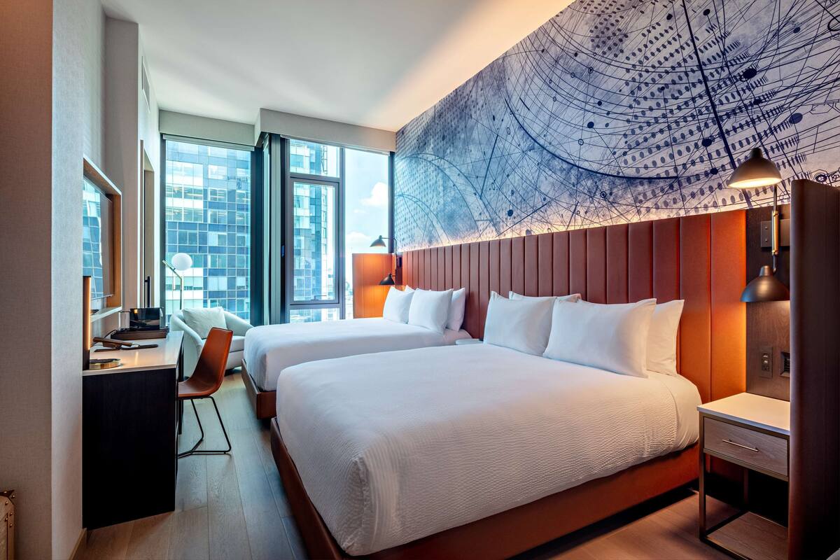 A hotel room with two beds, a desk with a chair, a large window, and a wall mural.