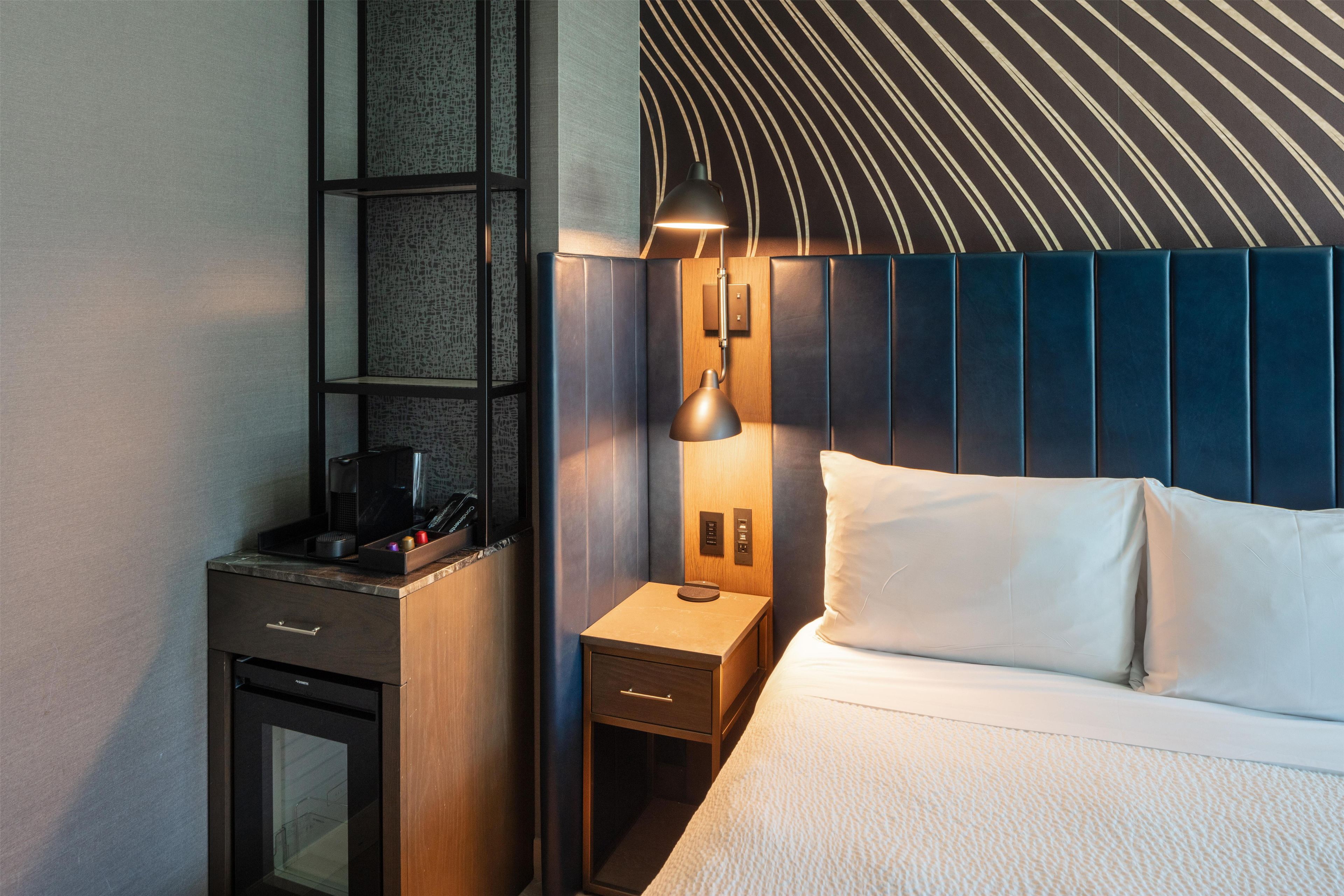 A hotel room with a bed, a nightstand, a lamp, a shelf with a coffee maker, and a wall with a textured pattern.