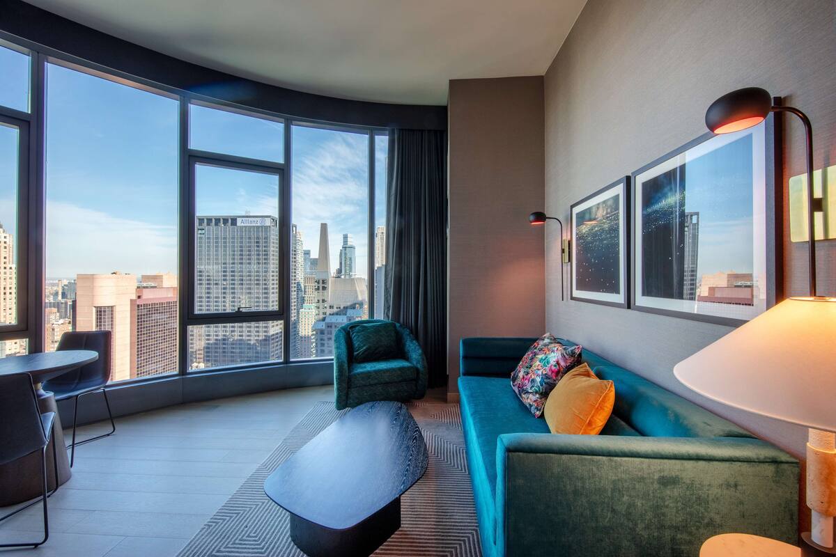 A modern hotel room with large windows offering a city view, a teal sofa, a small table, and a lamp.