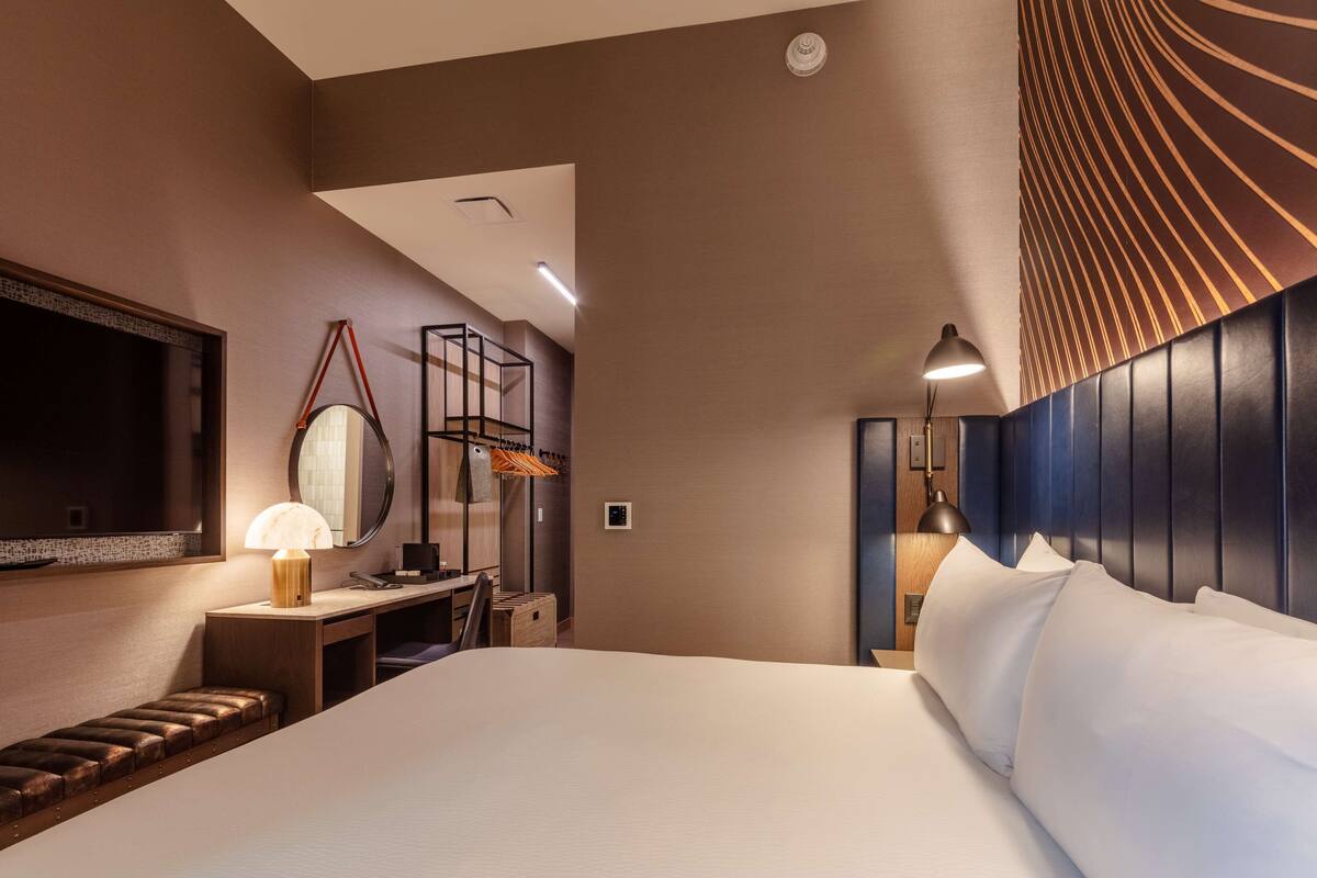 A hotel room with a large bed, a desk with a chair, a mirror, a lamp, and a wardrobe.