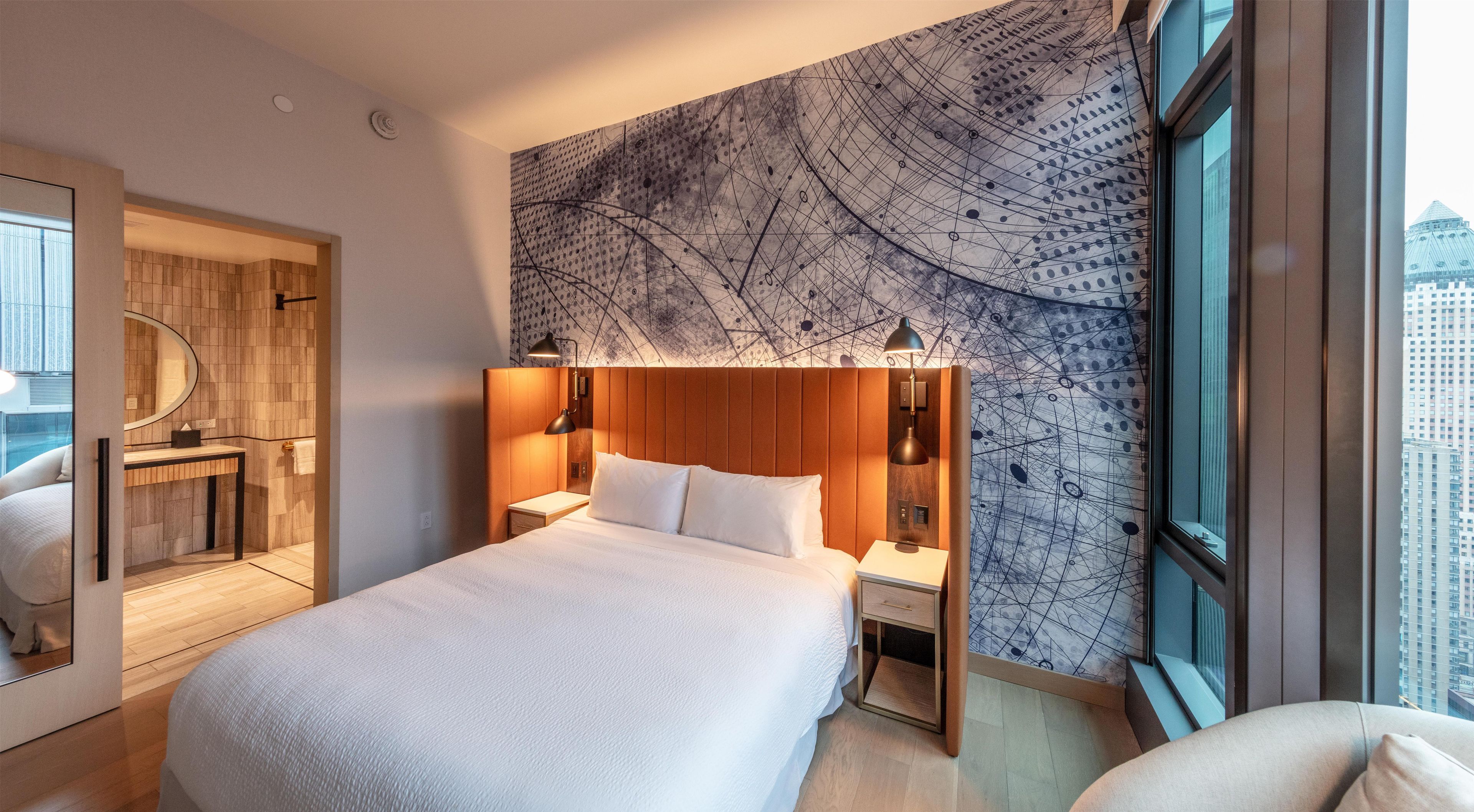 Una habitación de hotel moderna con una cama grande, cabecera de madera, mesita de noche, escritorio y un amplio ventanal con vista a la ciudad.