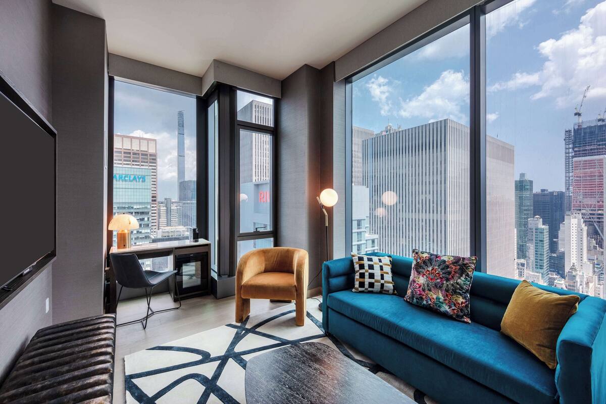 A modern hotel room with a large window offering a city view, a blue sofa, a wooden chair, and a desk with a lamp.