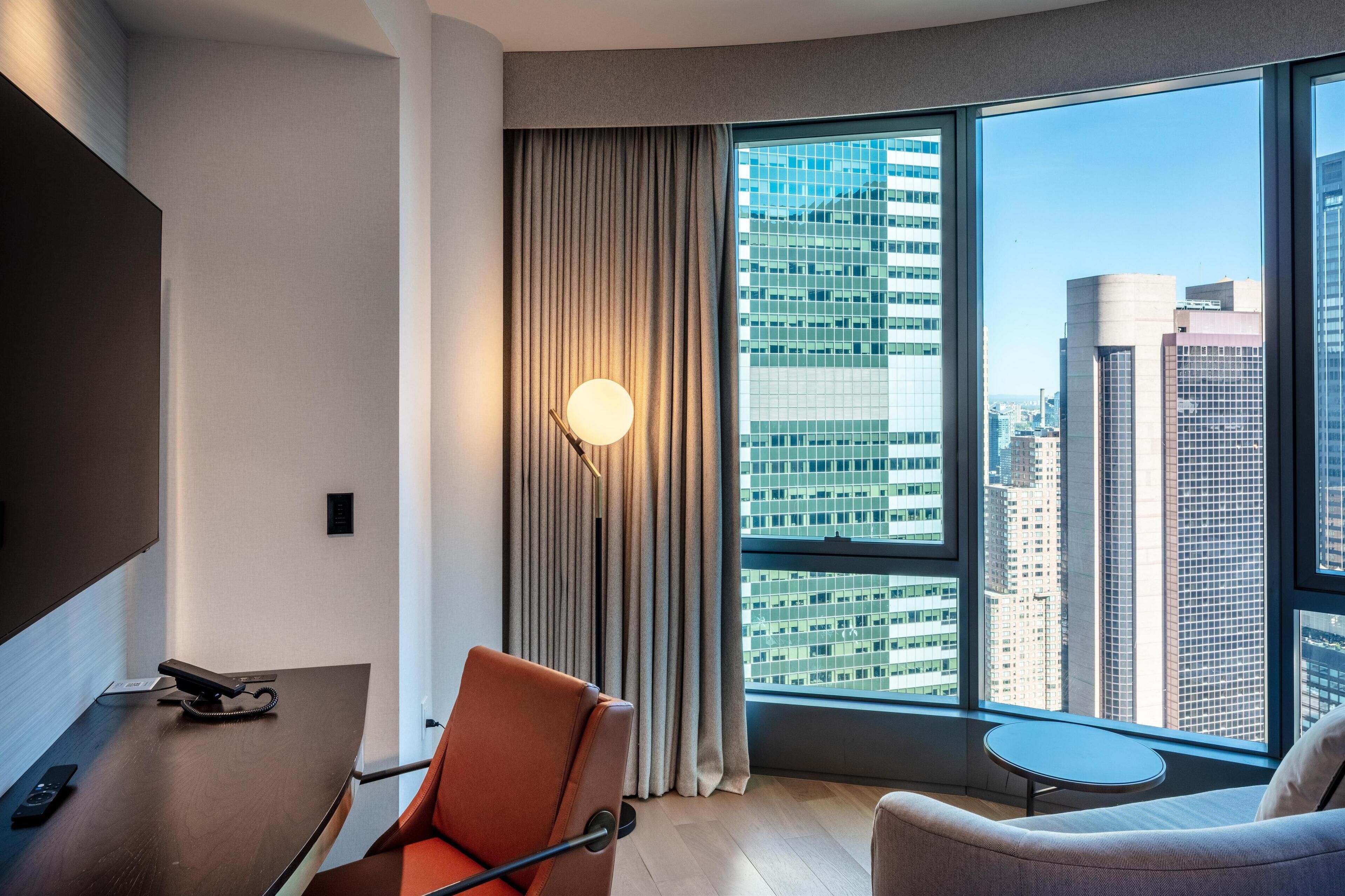 A modern hotel room with a large window offering a city view, a desk with a telephone, a chair, and a small table.