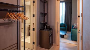 A hotel room with a wooden wardrobe, a black shelf unit, a small kitchen area, and a view of a green sofa and curtains in the adjacent room.
