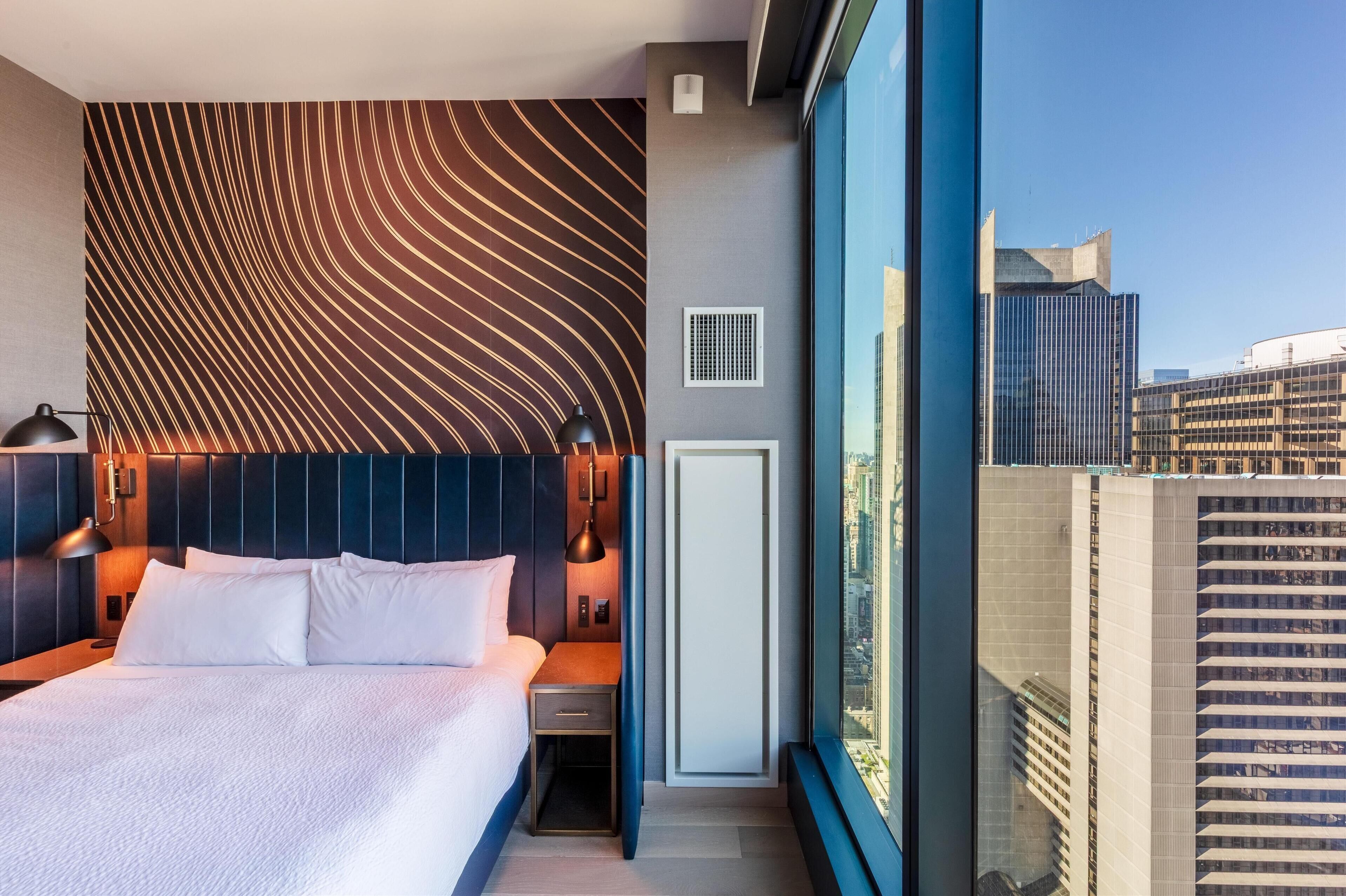 Un hotel moderno con una cama grande, un cabecero de diseño ondulado, una mesita de noche, una lámpara y vistas de la ciudad a través de amplios ventanales.