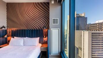 Un hotel moderno con una cama grande, un cabecero de diseño ondulado, una mesita de noche, una lámpara y vistas de la ciudad a través de amplios ventanales.