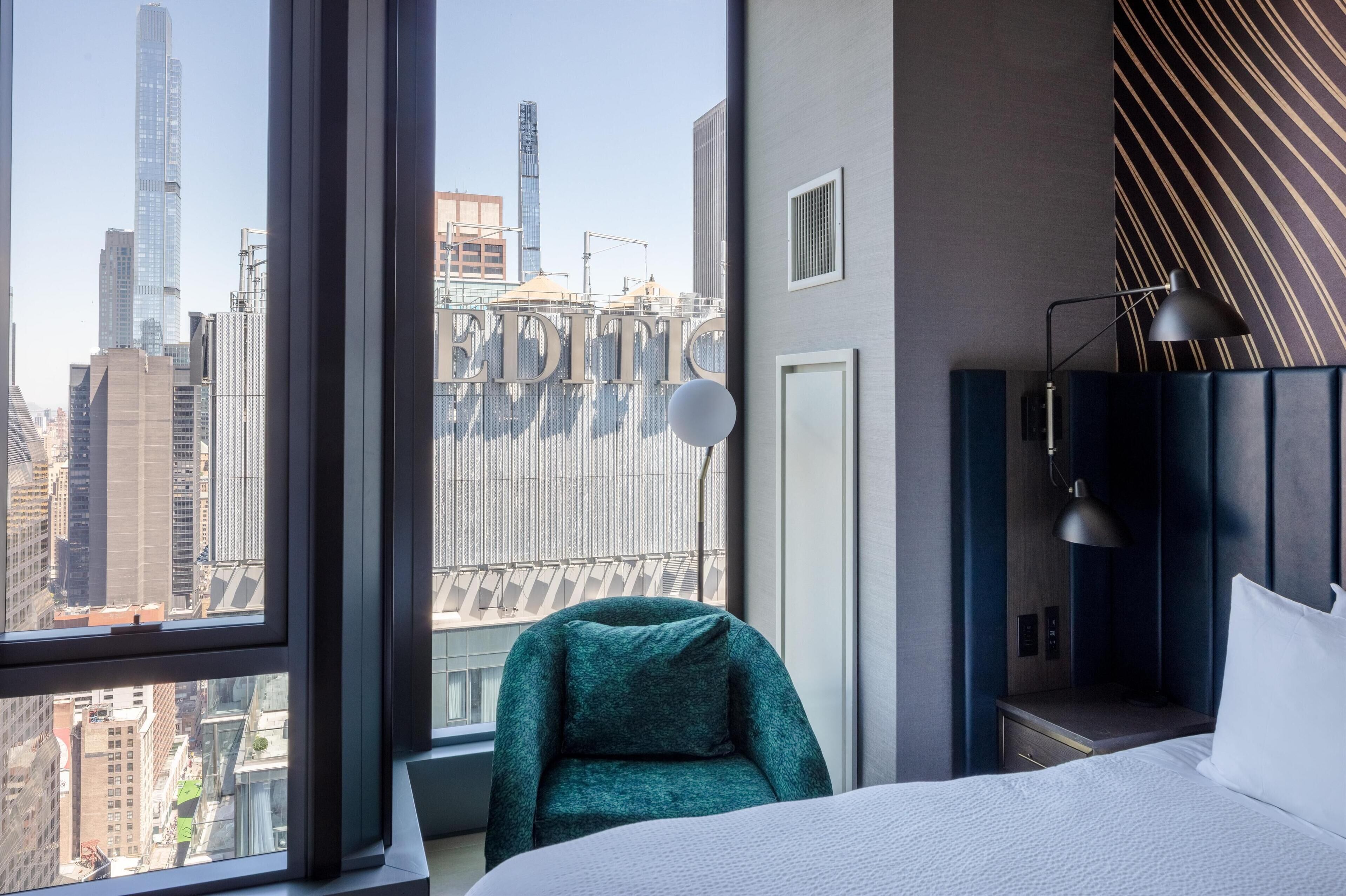 A modern hotel room with a large window offering a city view, a comfortable teal armchair, a neatly made bed, and a stylish lamp.