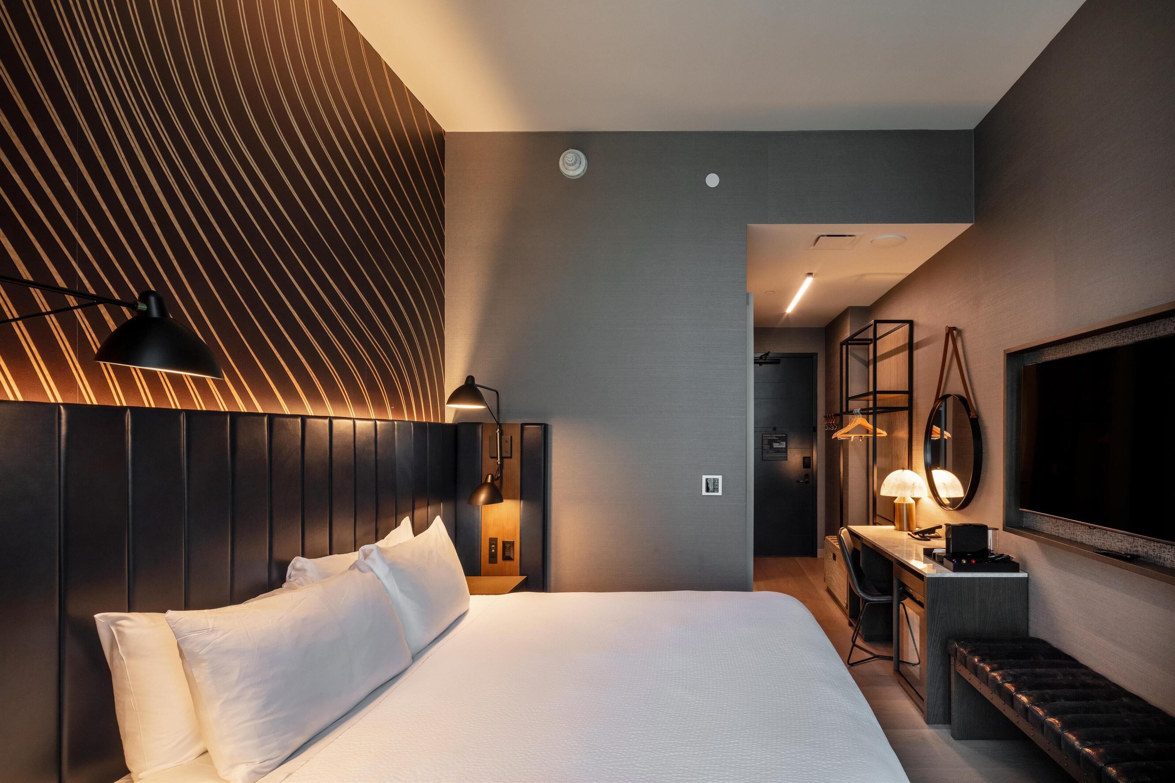 A modern hotel room with a large bed, bedside table, lamp, and a desk with a chair.