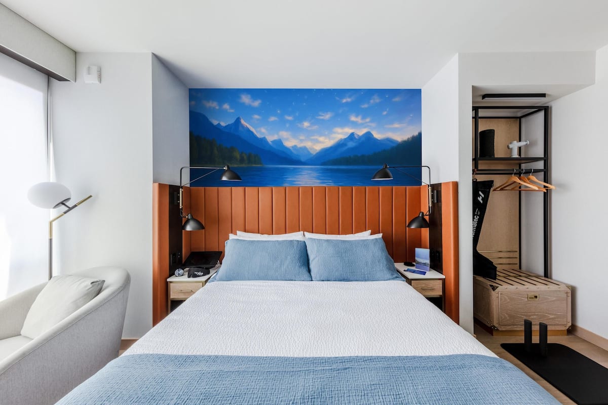 A bedroom with a bed, a nightstand, a lamp, a wardrobe, and a painting of a lake and mountains.