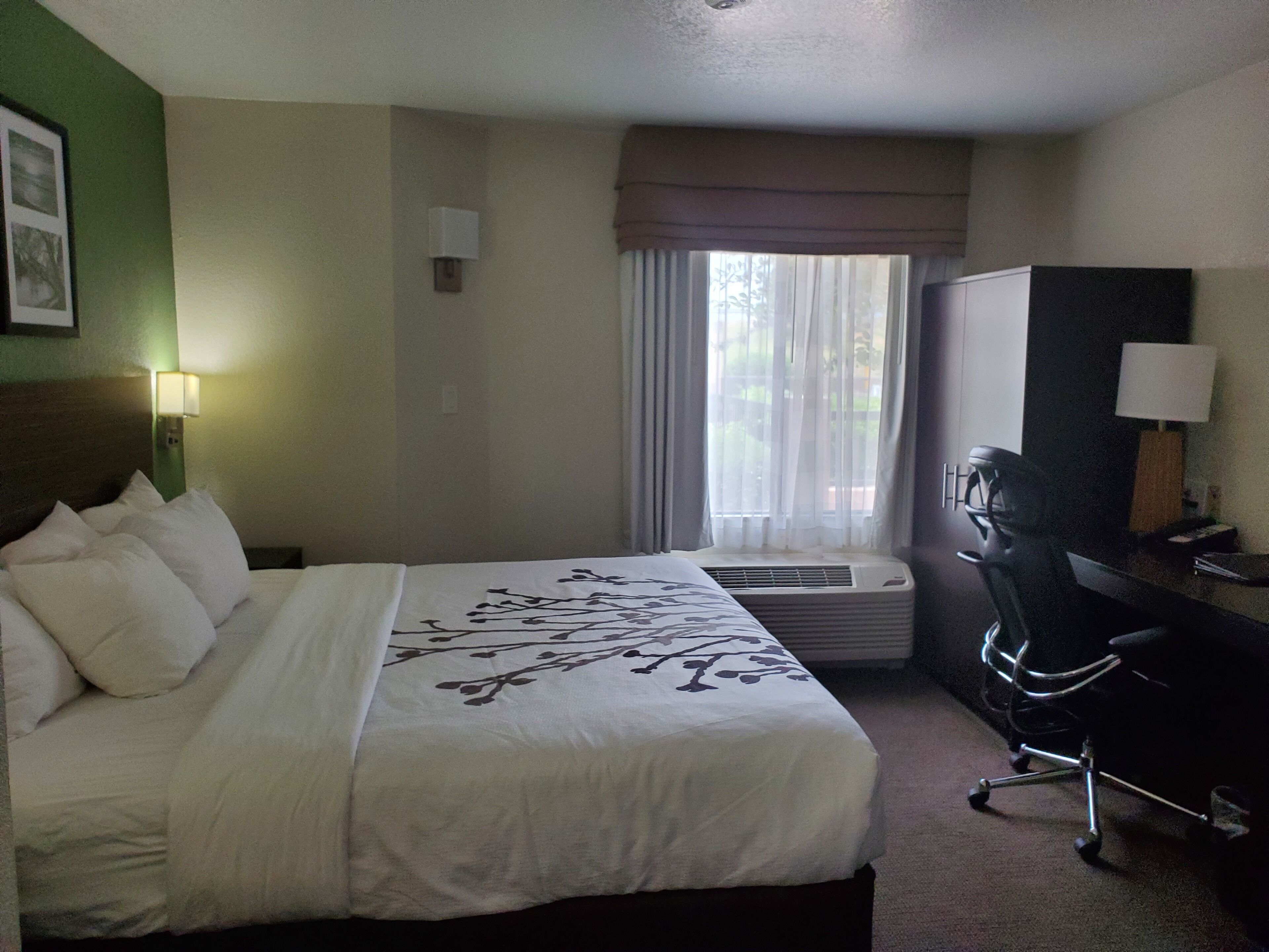 A hotel room with a bed, a desk, a chair, an air conditioning unit, and a window with curtains.
