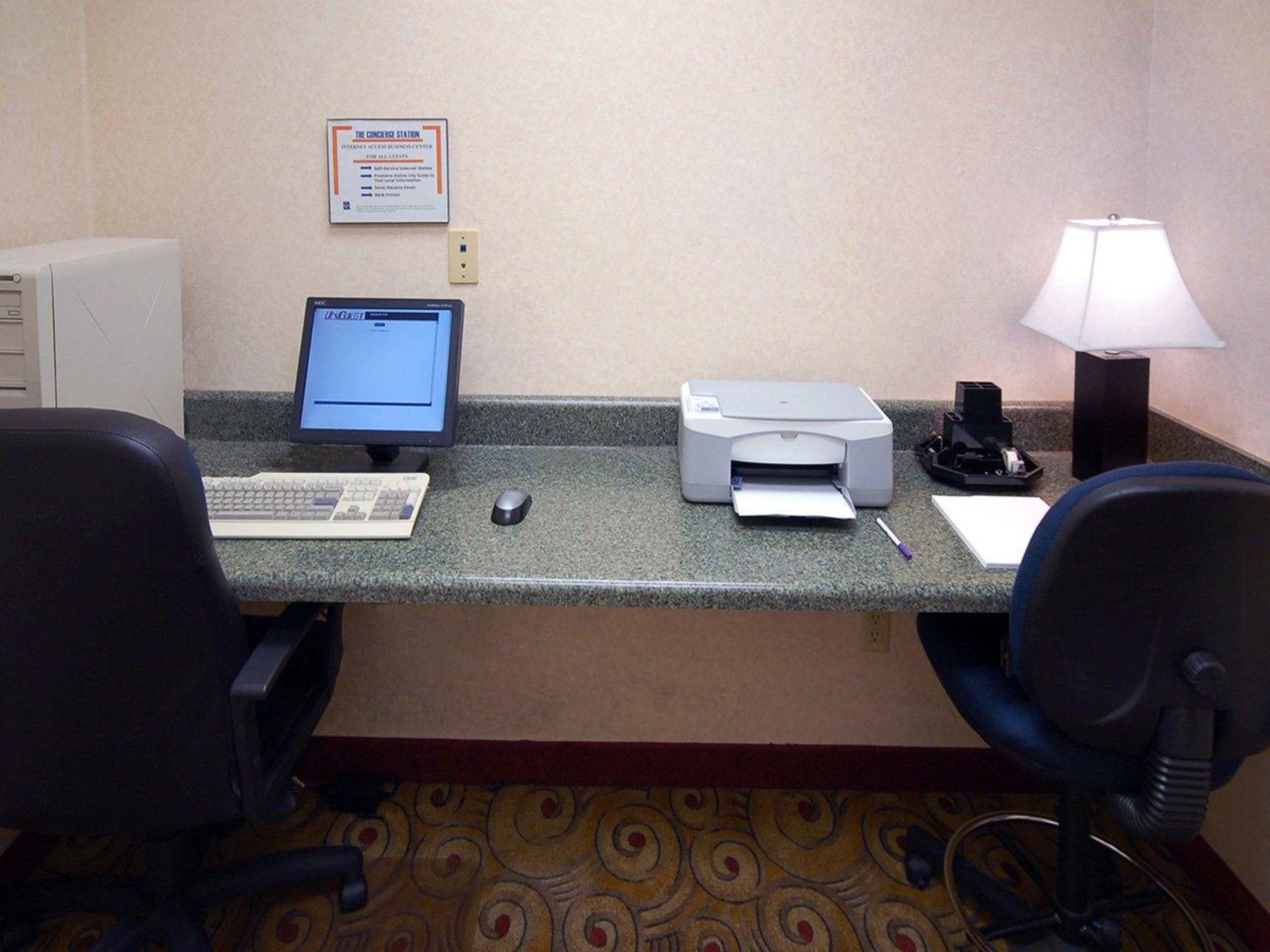 A hotel reception desk with a computer, printer, and desk lamp. There are two chairs and a carpeted floor.
