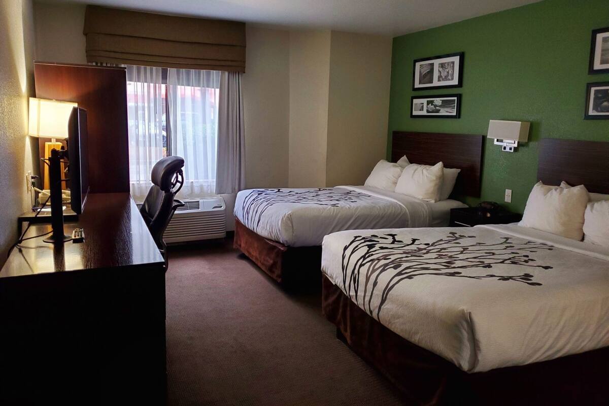 Ein Hotelzimmer mit zwei Betten, einem Schreibtisch mit Computer, einem Stuhl und einem Vorhangfenster.