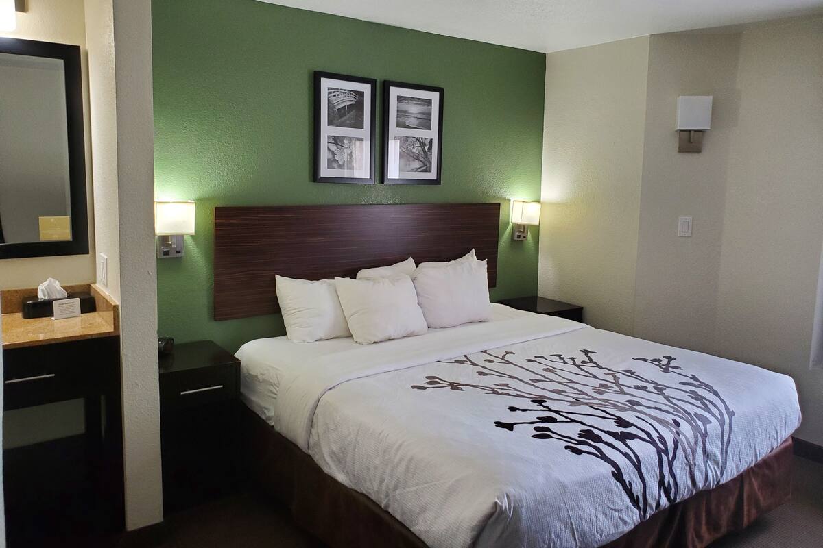 Ein ordentlich gemachtes Bett mit weißer Decke und Kissen, ein hölzernes Kopfende, zwei gerahmte Bilder an der Wand und ein Nachttisch mit einer Lampe.