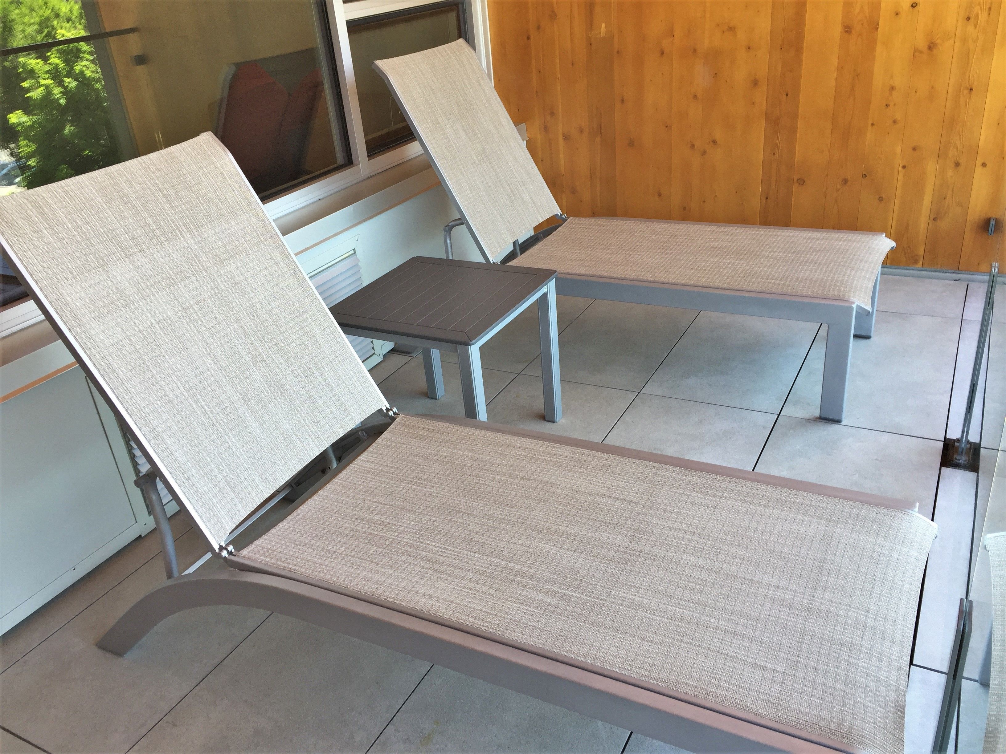 A room with two reclining chairs with fabric seats and metal frames, a wooden wall, and a window with blinds.
