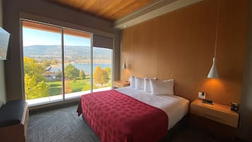 A bedroom with a large window, a bed with a red polka dot bedspread, a wooden headboard, a bedside table with a lamp, and a view of a lake and hills.
