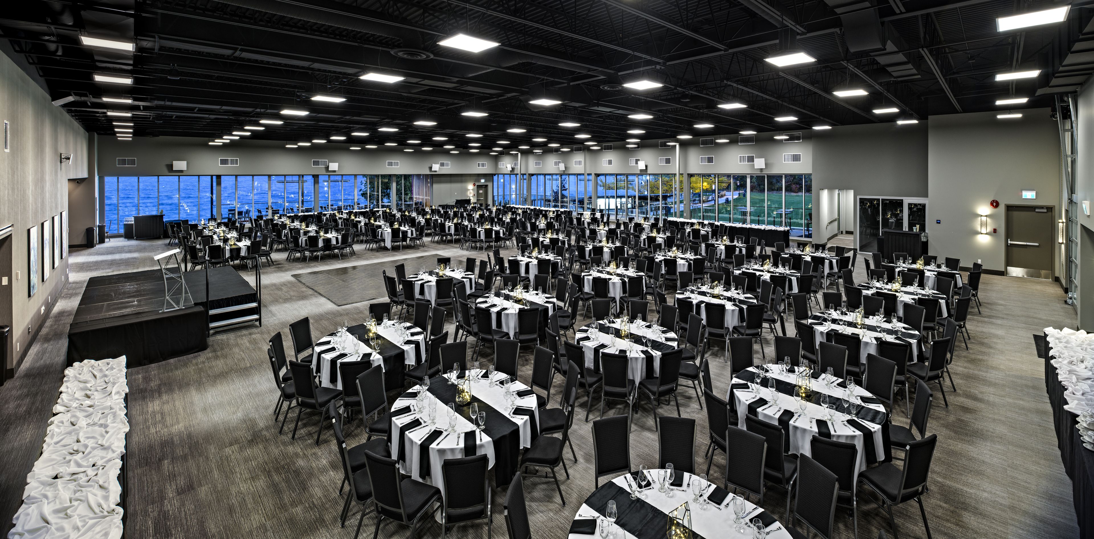 Un salón de conferencias amplio con varias mesas dispuestas para comer, un escenario y ventanas grandes que ofrecen vistas al exterior.