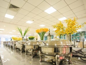 Una larga fila de carros buffet plateados en una habitación luminosa y amplia, con techo de baldosas y grandes ventanales que dejan entrar luz natural.