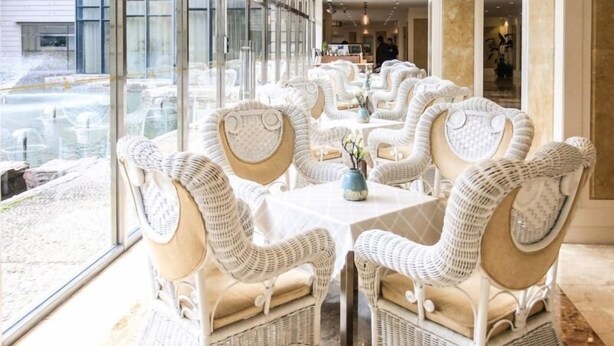 A restaurant with wicker chairs and tables, large windows, and a view of the street.