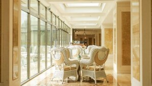 A spacious, well-lit hallway with a high ceiling and large windows, leading to a dining area with wicker furniture and a polished floor.