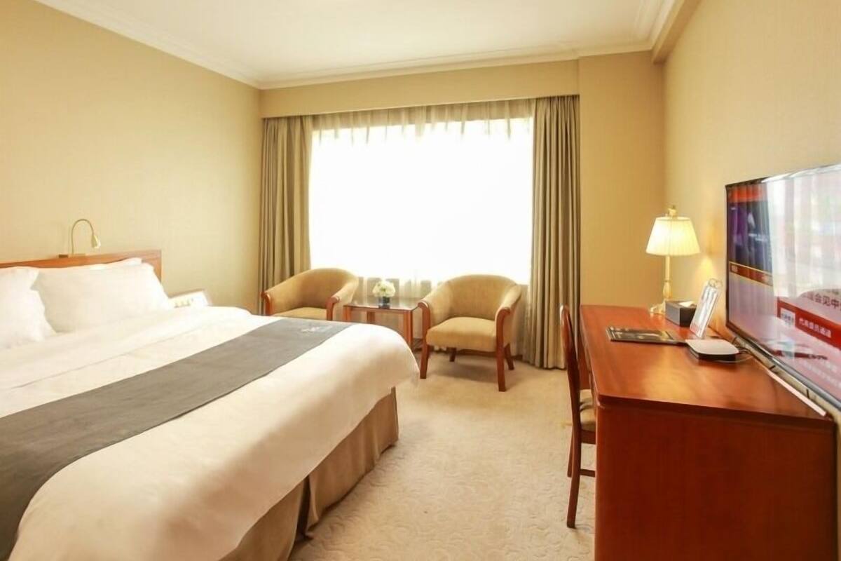 A hotel room with a bed, two armchairs, a desk with a lamp, a television, and a window with curtains.