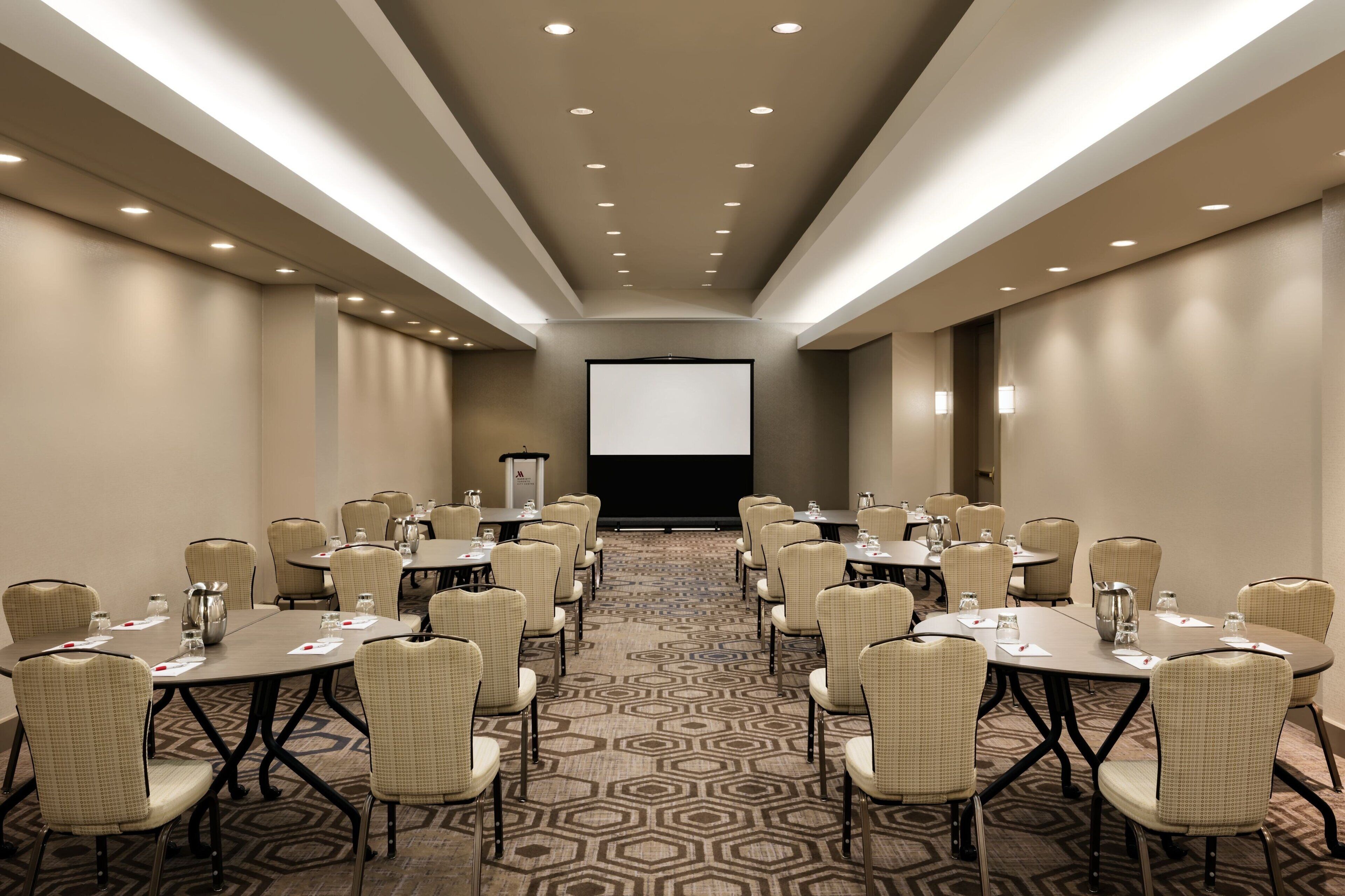 Una sala conferenze con diversi tavoli e sedie, uno schermo per proiettore e un pavimento rivestito di moquette.