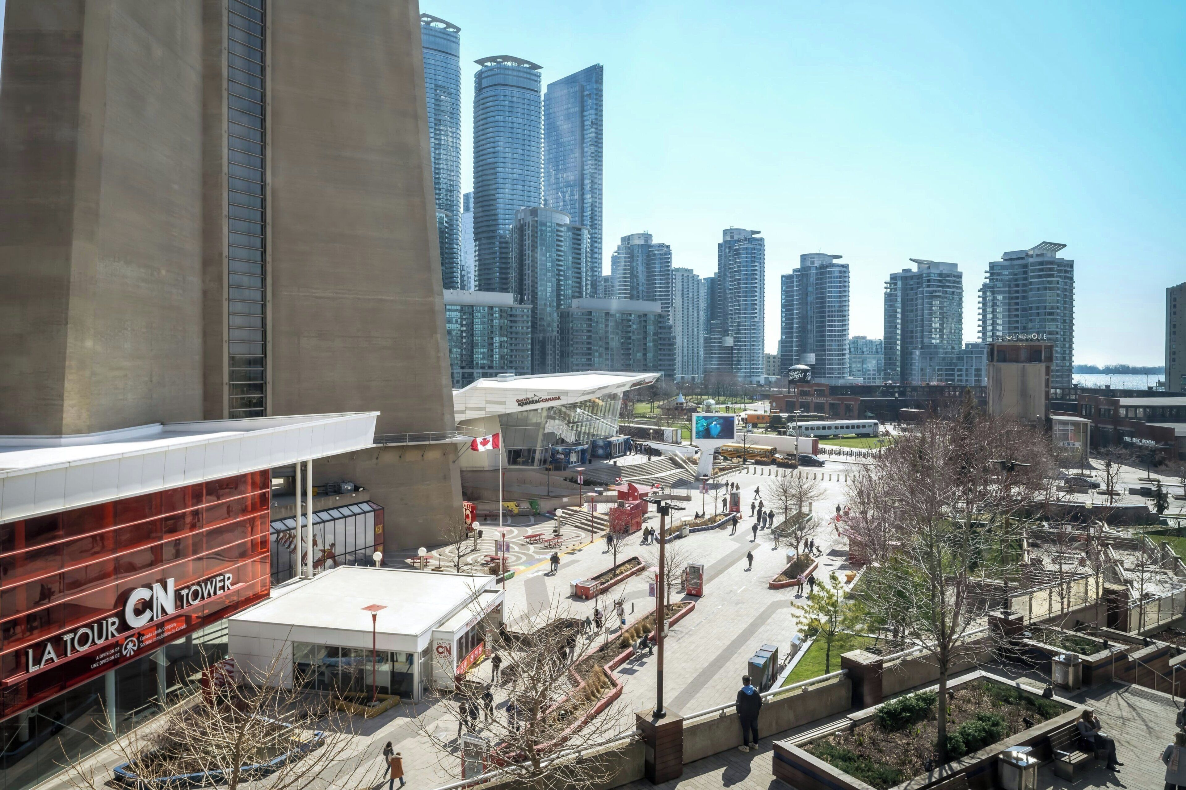 Et moderne bytorv med høje bygninger, en markant rød bygning med skiltet 'LA TOUR CN TOWER', og en klar blå himmel.