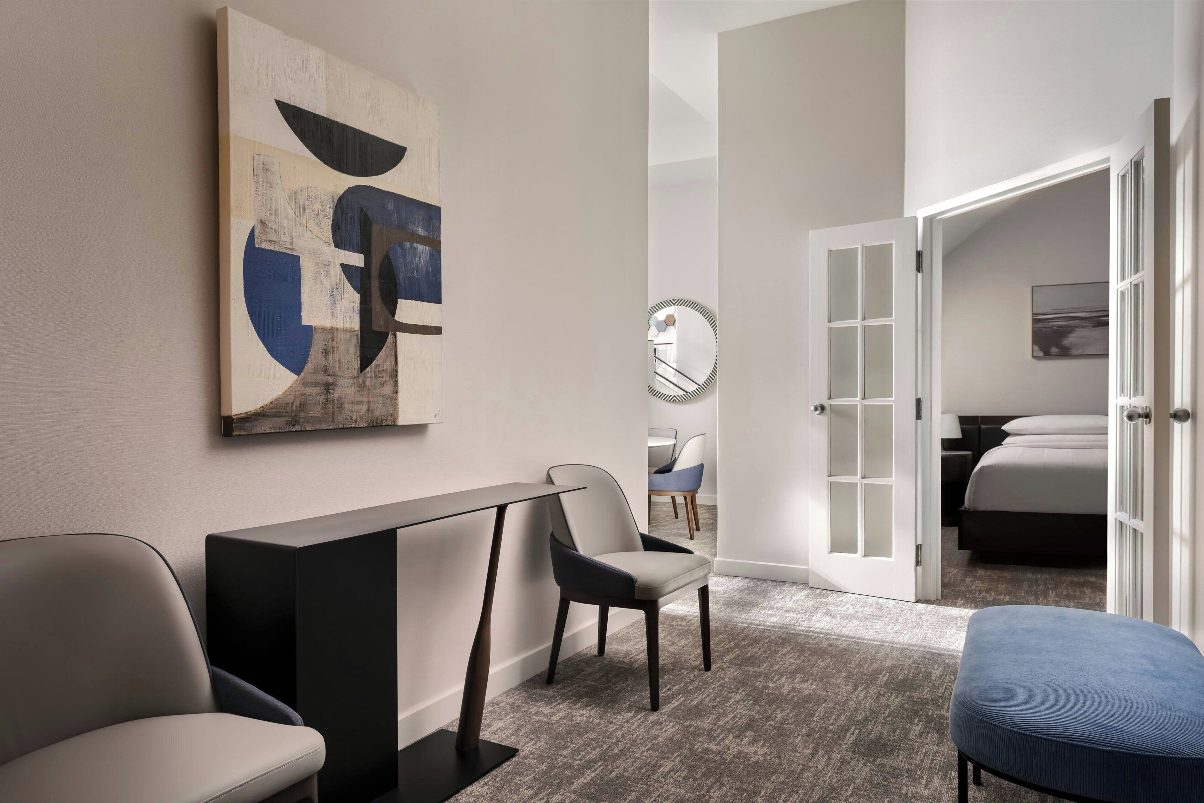 Et hotelværelse med moderne design, med et stort abstrakt maleri, et spisebord, to stole, et spejl og udsigt til et soveværelse med en seng og et natbord.