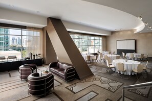 Una habitación de hotel moderna con un ventanal grande, un sofá, un área de comedor con mesas redondas y una pantalla para proyector.