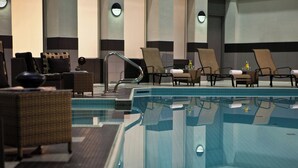 An indoor swimming pool area with lounge chairs, a small table with a bottle, and a chair with a towel.