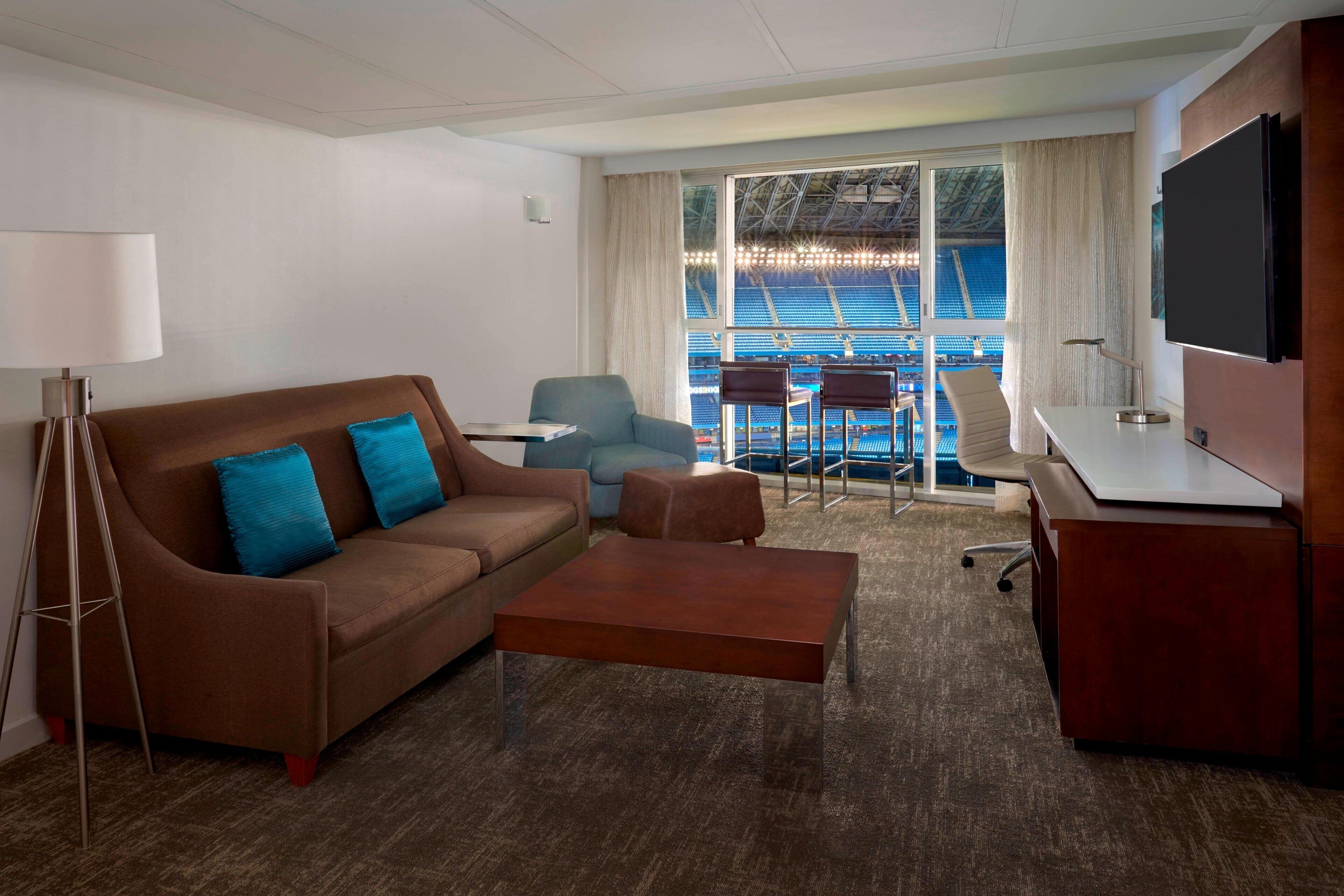 Habitación de hotel con un sofá marrón, una mesita, un escritorio, un televisor y una ventana con vista a un estadio.
