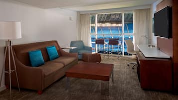 Habitación de hotel con un sofá marrón, una mesita, un escritorio, un televisor y una ventana con vista a un estadio.