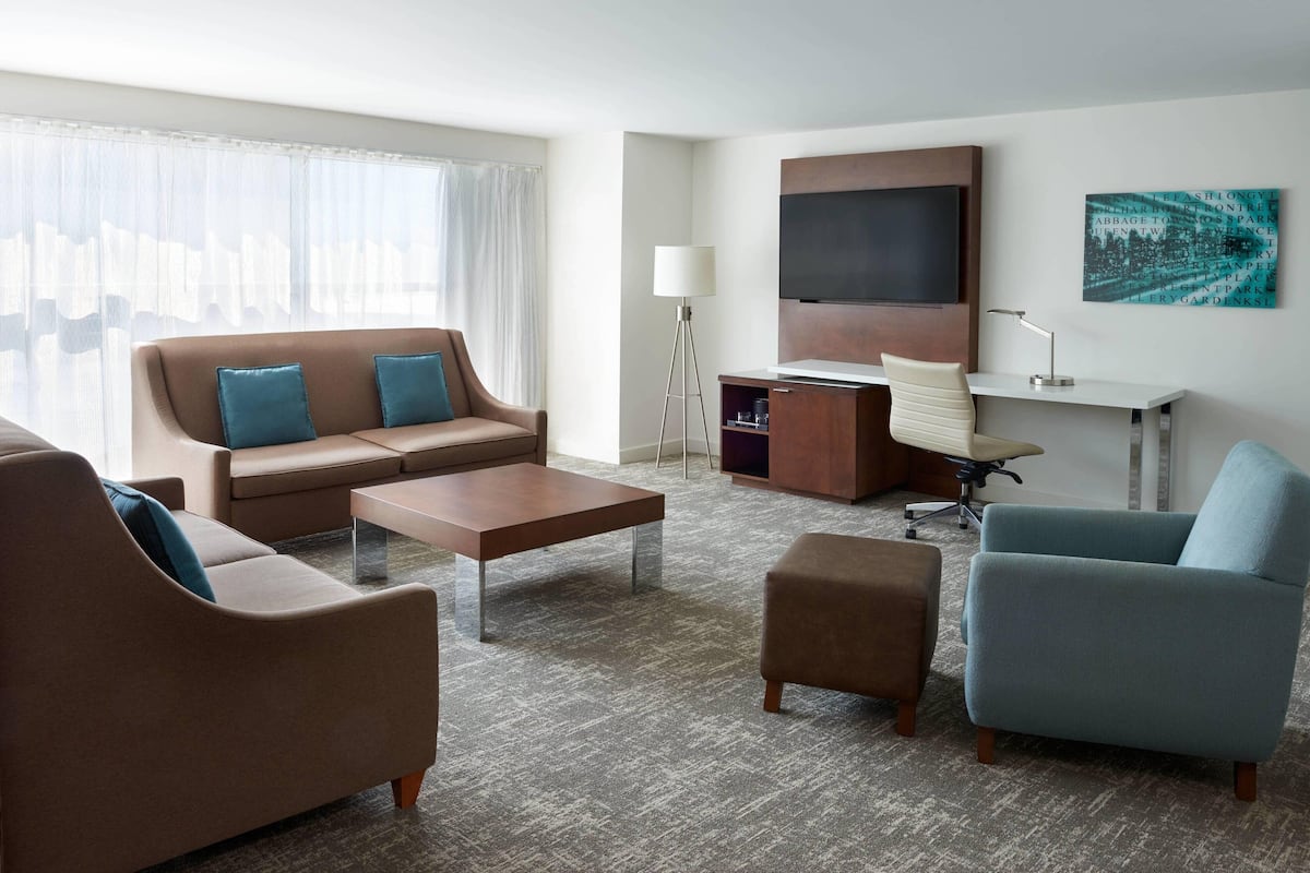Et moderne hotelværelse med en sofa, et sofabord, et skrivebord med stol, et fjernsyn og et billede på væggen.