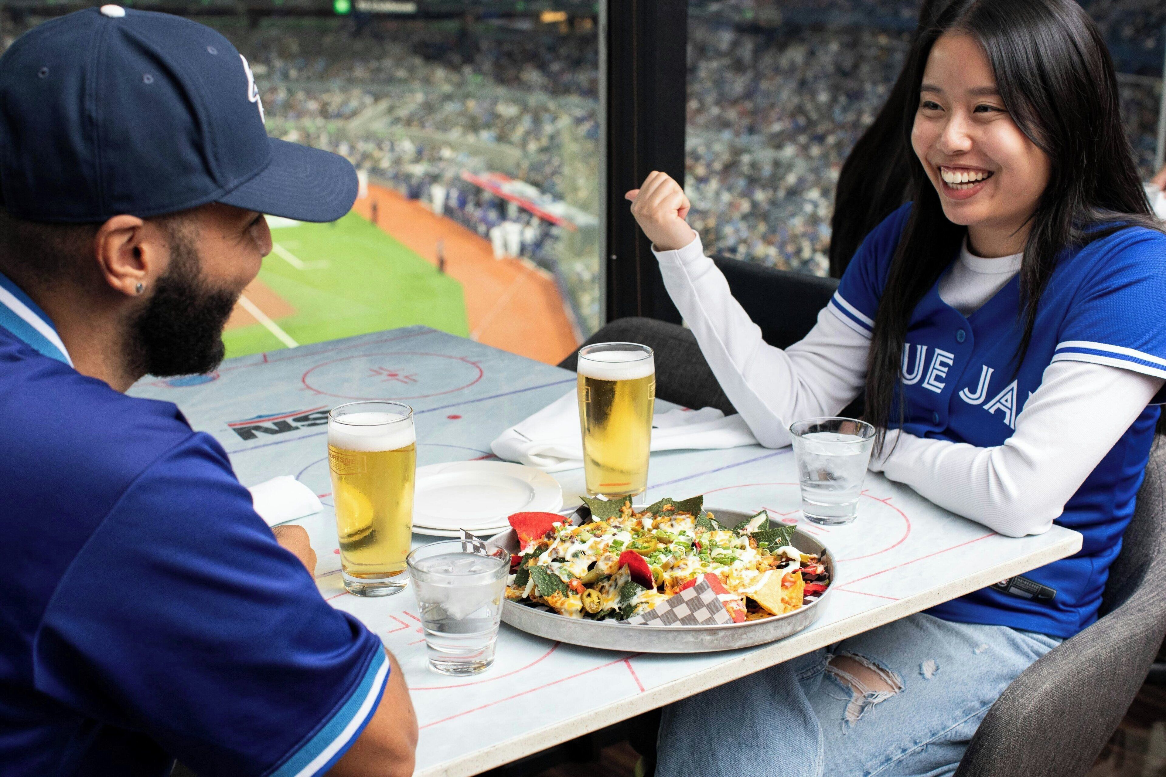 Uma mesa de jantar com refeição e bebidas, vista para um campo de beisebol através de uma janela, e uma pessoa usando boné e camisa azuis.