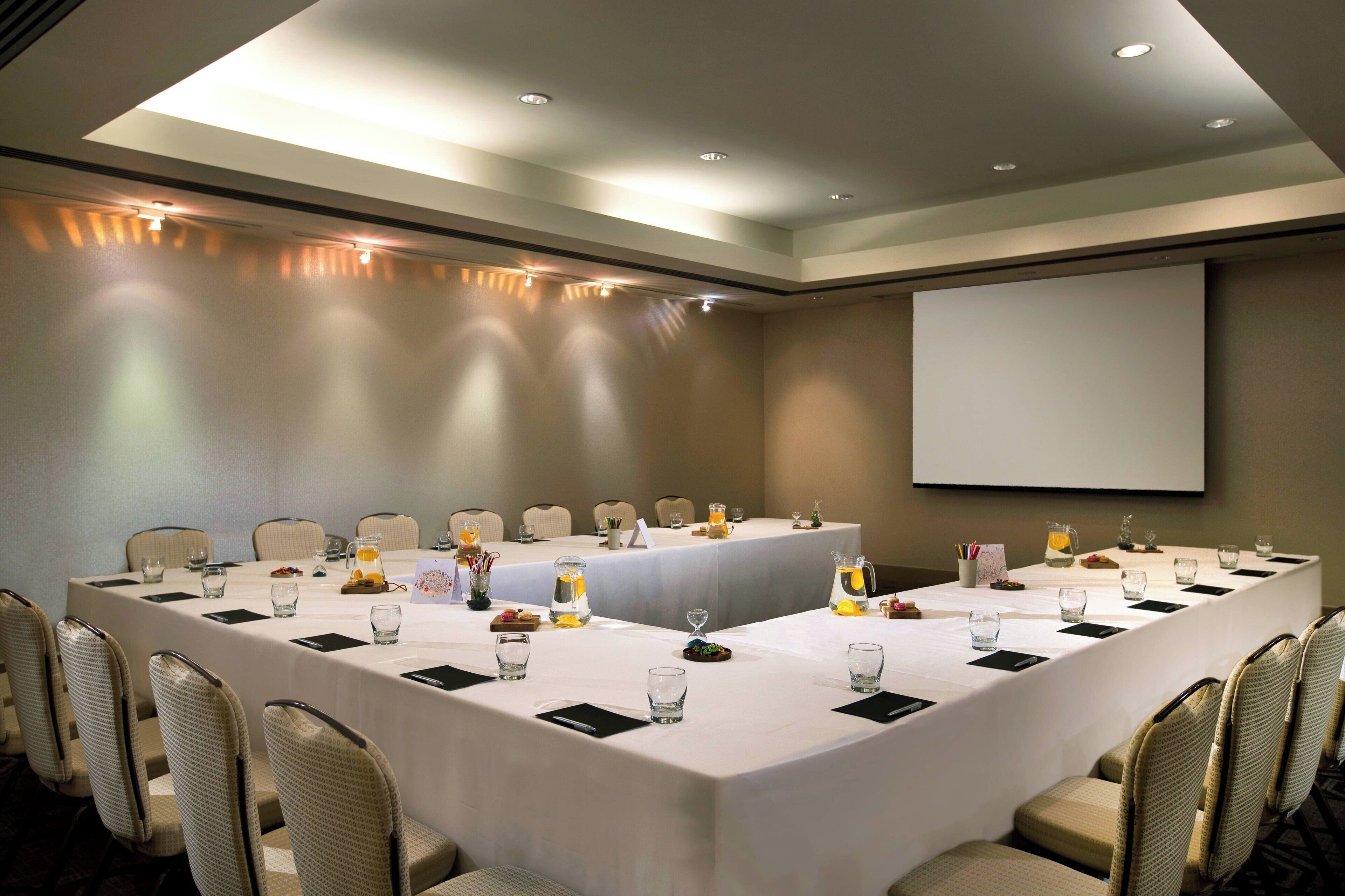 Una sala de conferencias con una larga mesa, sillas, una pantalla de proyector y una pizarra.
