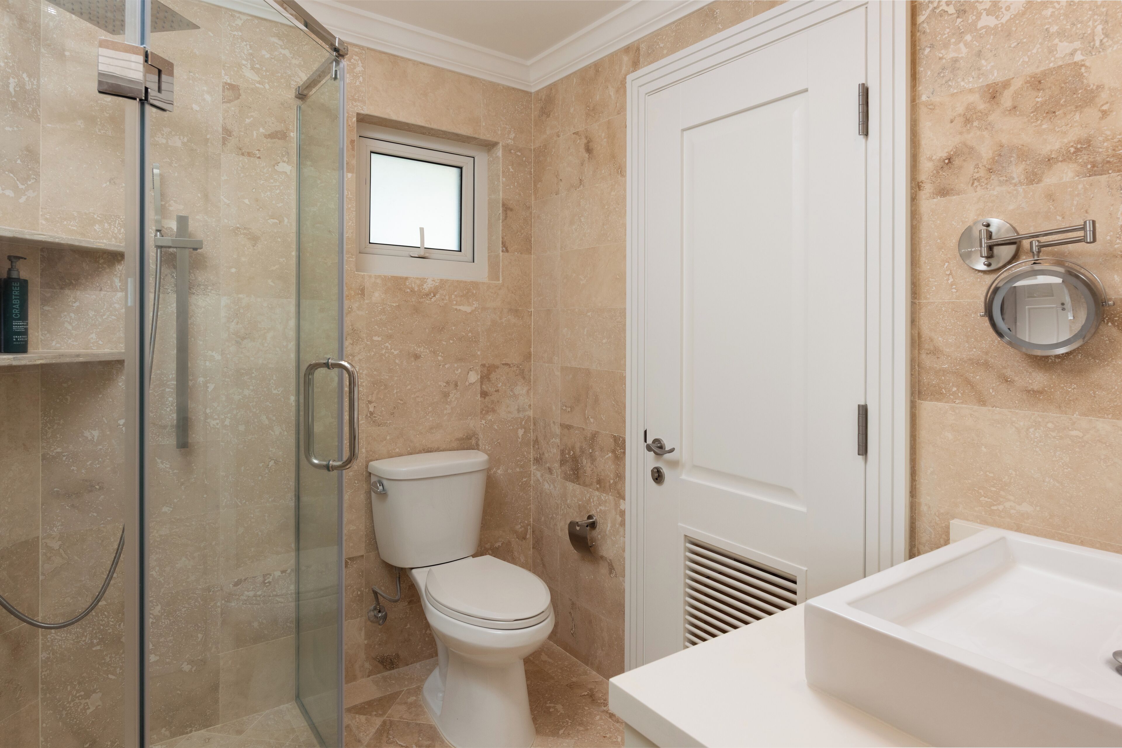 A bathroom with a glass-enclosed shower, a toilet, a sink, and a window above the toilet.
