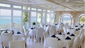 Un restaurant avec des tables et des chaises blanches, de grandes fenêtres donnant sur l’océan et des ventilateurs de plafond.