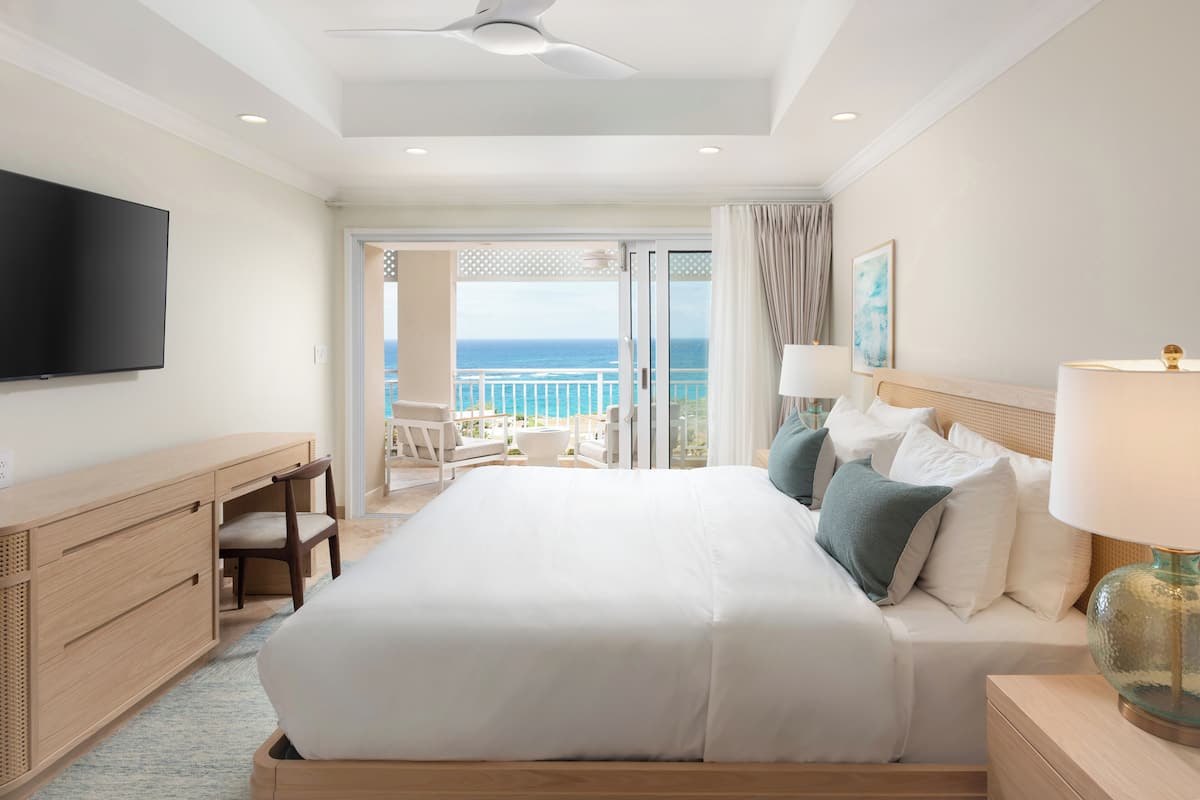 Habitación de hotel con una cama grande, un escritorio, una silla, un televisor y un balcón con vista al océano.