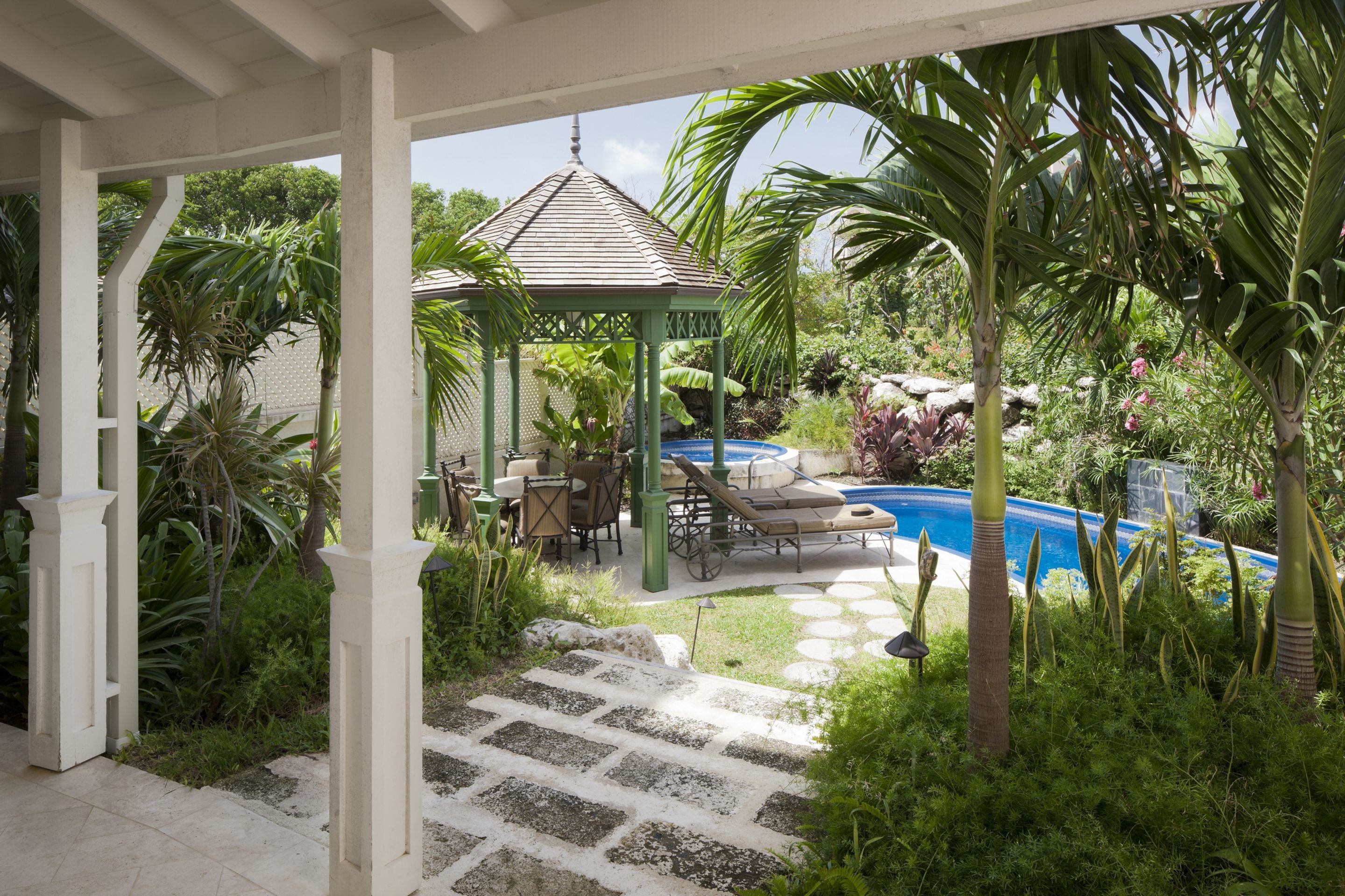 A covered outdoor area with a gazebo, lounge chairs, and a swimming pool surrounded by tropical plants.