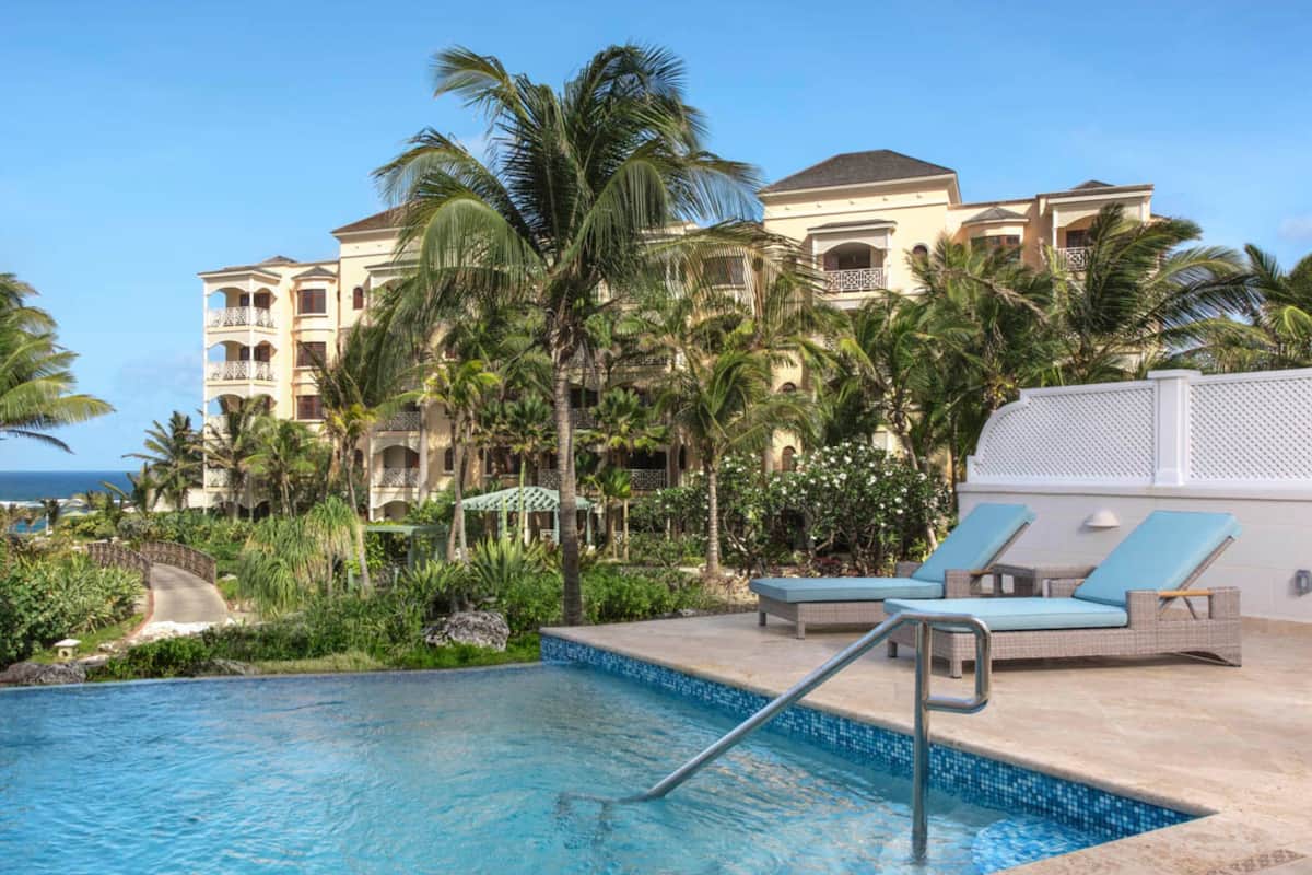 Zona de piscina con sillones de descanso, palmeras y un edificio con balcones al fondo.
