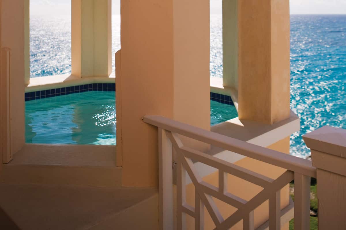 Un área de piscina con agua azul cristalina, rodeada de muros y columnas color beige, con vista al océano.
