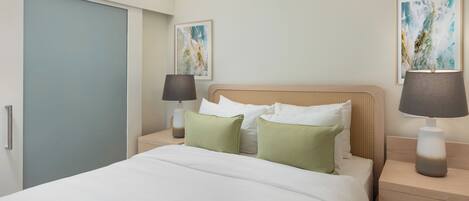 Quarto de hotel com uma cama, duas mesas de cabeceira, um criado-mudo, uma luminária e duas molduras na parede.