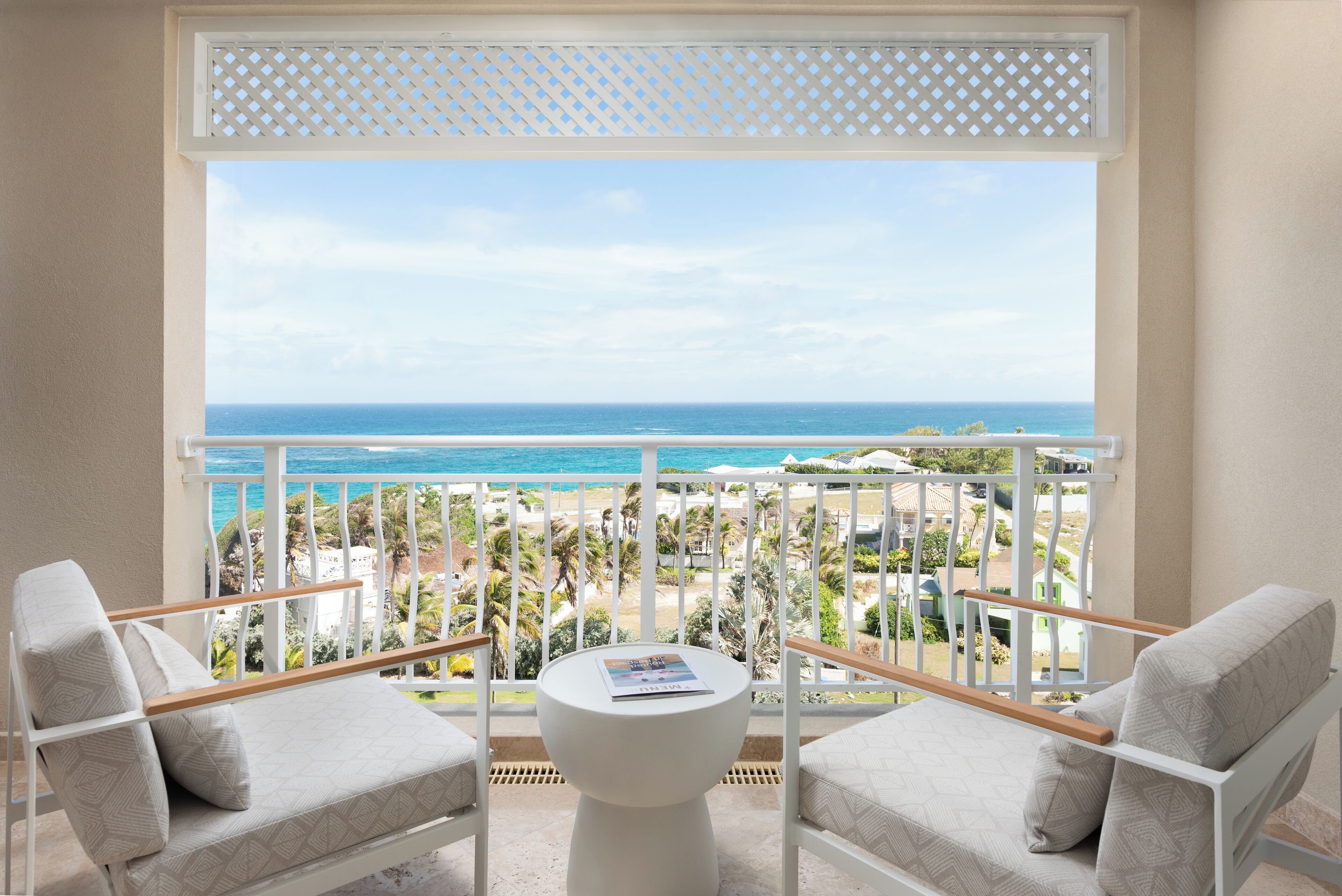A balcony with a view of the ocean, two chairs, a small table, and a white lattice window blind.