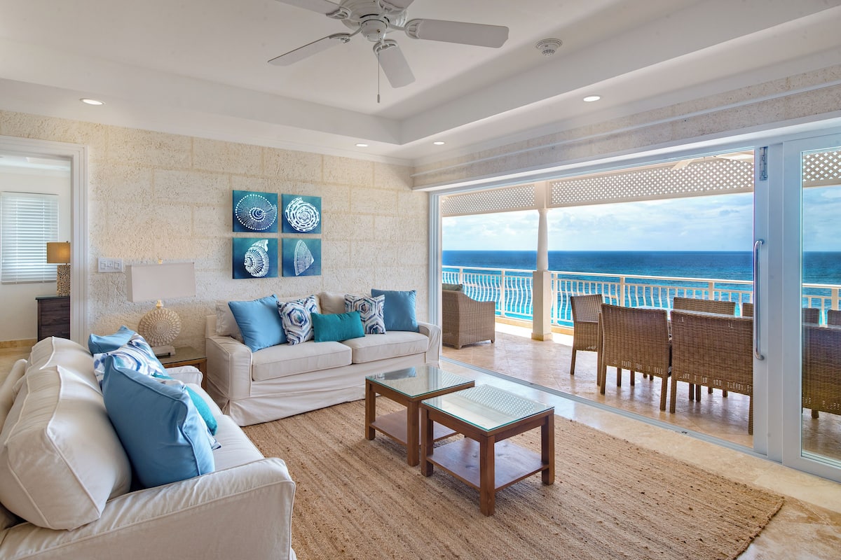 Amplia sala de estar con un sofá blanco, cojines azules y una mesa de centro de vidrio. Se observan dos obras de arte en la pared y un ventilador de techo. La habitación cuenta con un gran ventanal corredero que da a un balcón con mobiliario de mimbre y vista al océano.