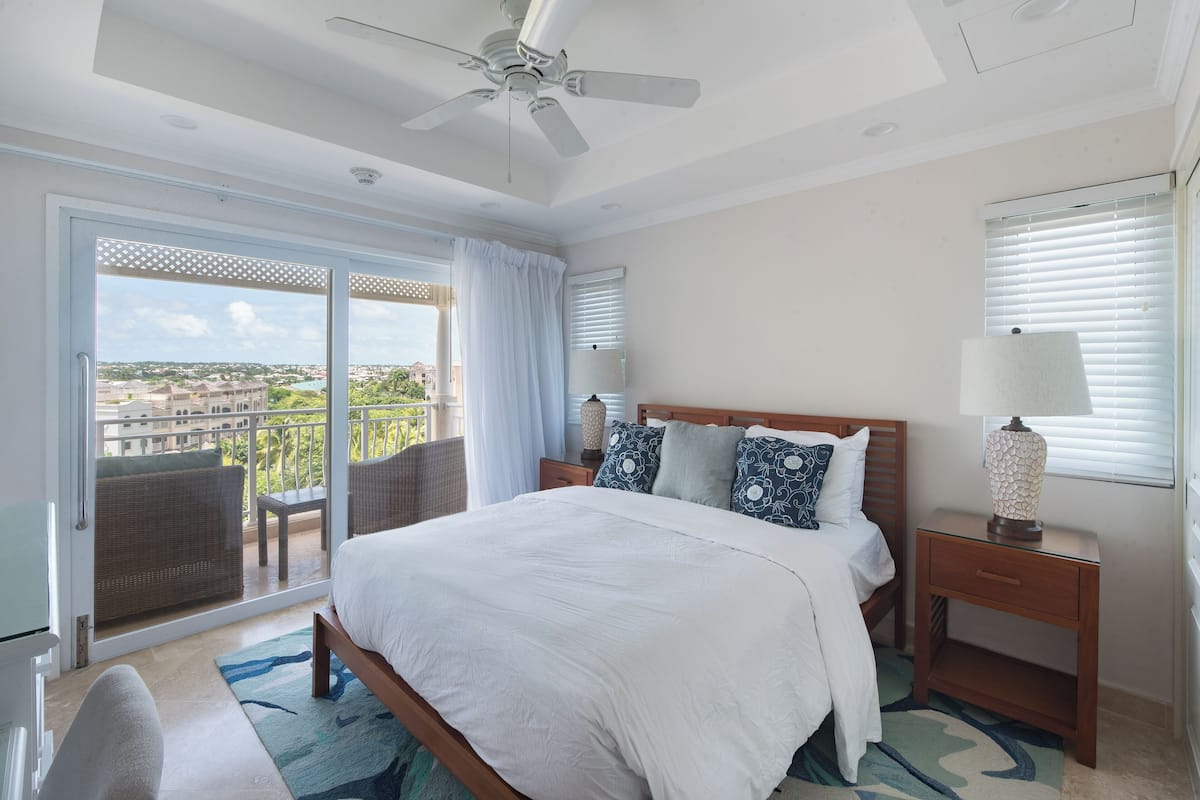 Habitación de hotel con cama, mesita de noche, lámpara, silla y vista a la ciudad a través de amplias ventanas.