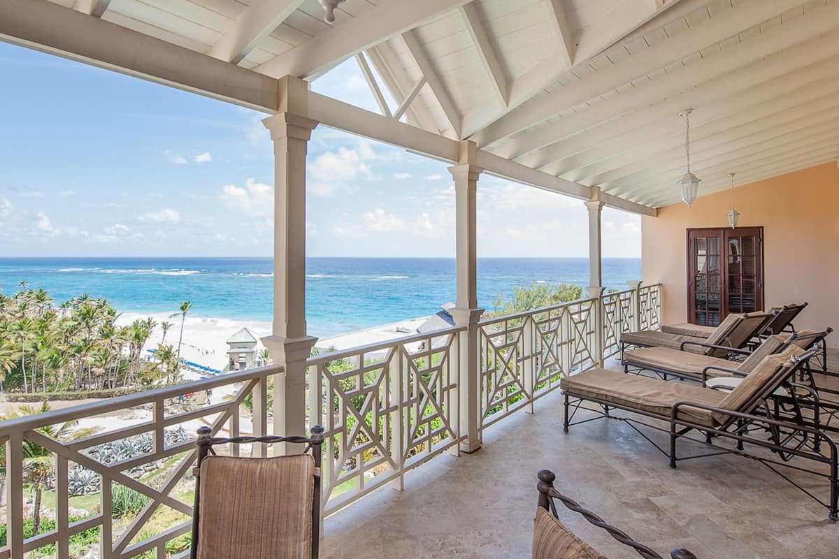 Un balcón techado con vistas al océano y la playa, amueblado con sillones y una pequeña mesa.