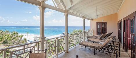 Un balcón techado con vistas al océano y la playa, amueblado con sillones y una pequeña mesa.