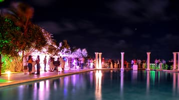 Zona de piscina iluminada de noche con columnas y varias personas.