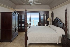 A bedroom with a four-poster bed, a wooden wardrobe, and a view of the ocean through a balcony with shutters.